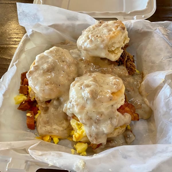 Kono's biscuits and gravy with scrambled eggs, bacon, and spam in a take-out container.