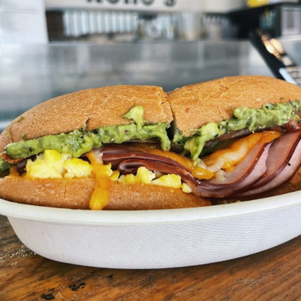 A Kono's breakfast sandwich with scrambled eggs, ham, cheese, and guacamole in a white bowl.