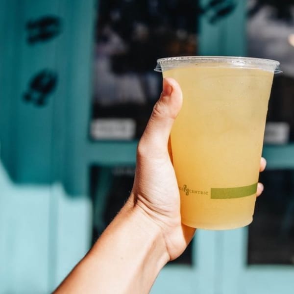 A hand holds a clear plastic cup of Kono's Hawaiian lemonade against a blue wall.