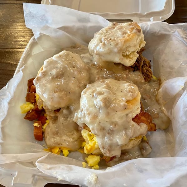 Kono's biscuits and gravy with scrambled eggs, bacon, and potatoes in a takeout container.