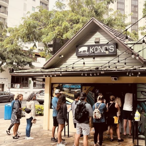 People queue outside Kono's, a Hawaiian restaurant with a pig logo and string lights.
