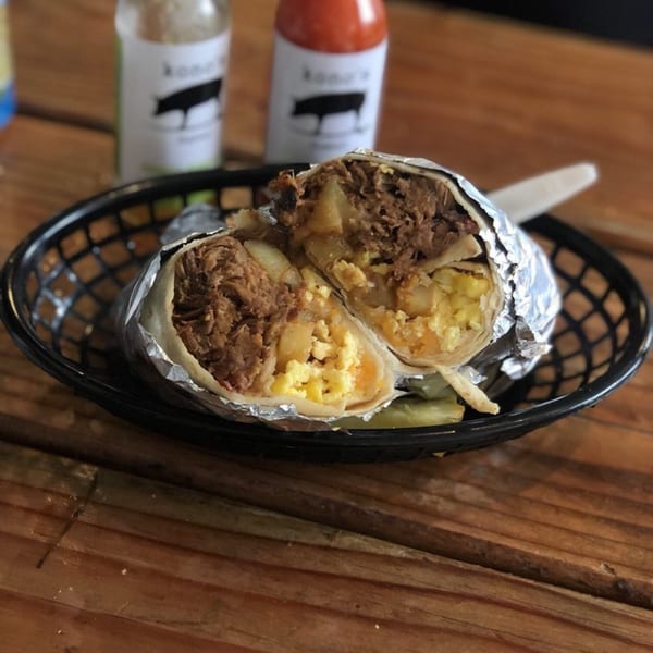 Kono's breakfast burrito, cut in half, revealing scrambled eggs, potatoes, and pulled pork, with hot sauces in background.